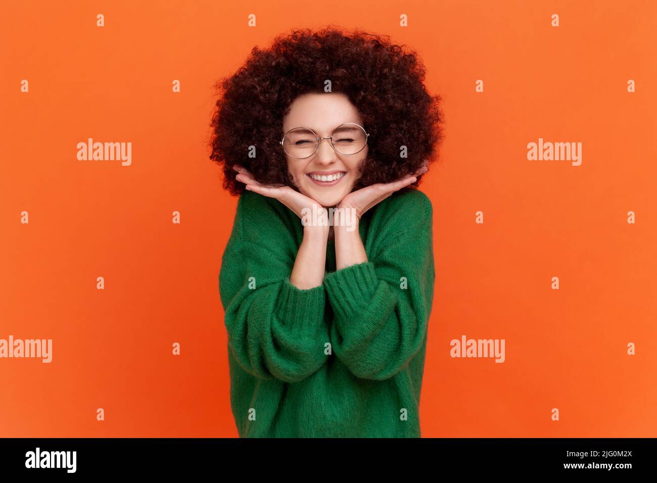 Happy woman with Afro hairstyle wearing green casual style sweater standing with hands under chin, keeping eyes closing, expressing happiness. Indoor studio shot isolated on orange background. Stock Photo