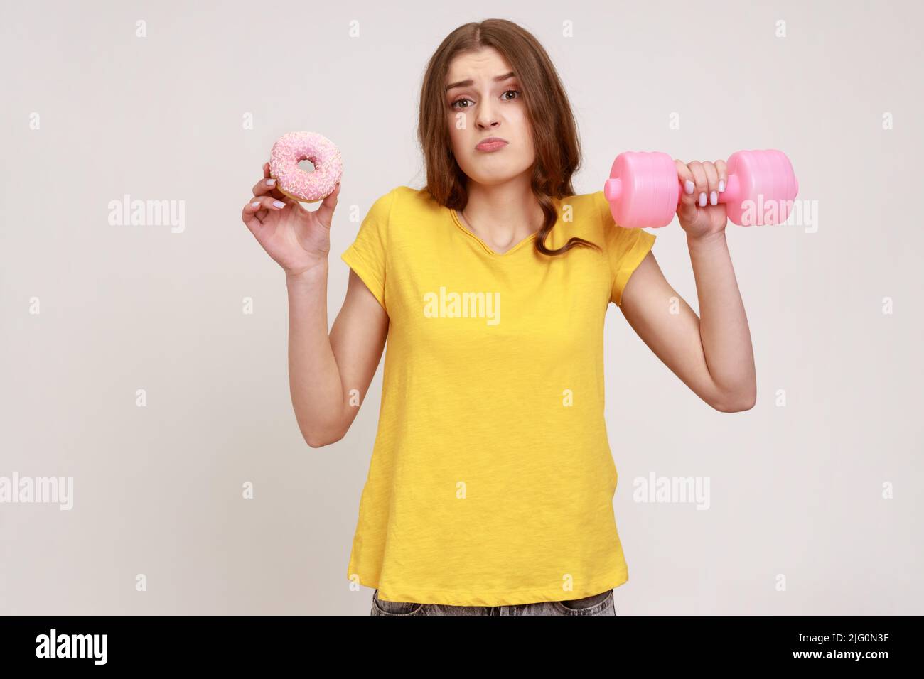 Confused teenager girl in yellow casual style T-shirt holding in hands pink dumbbell and donut with pink glaze, making hard choice, succumb to seduction. Indoor studio shot isolated on gray background Stock Photo