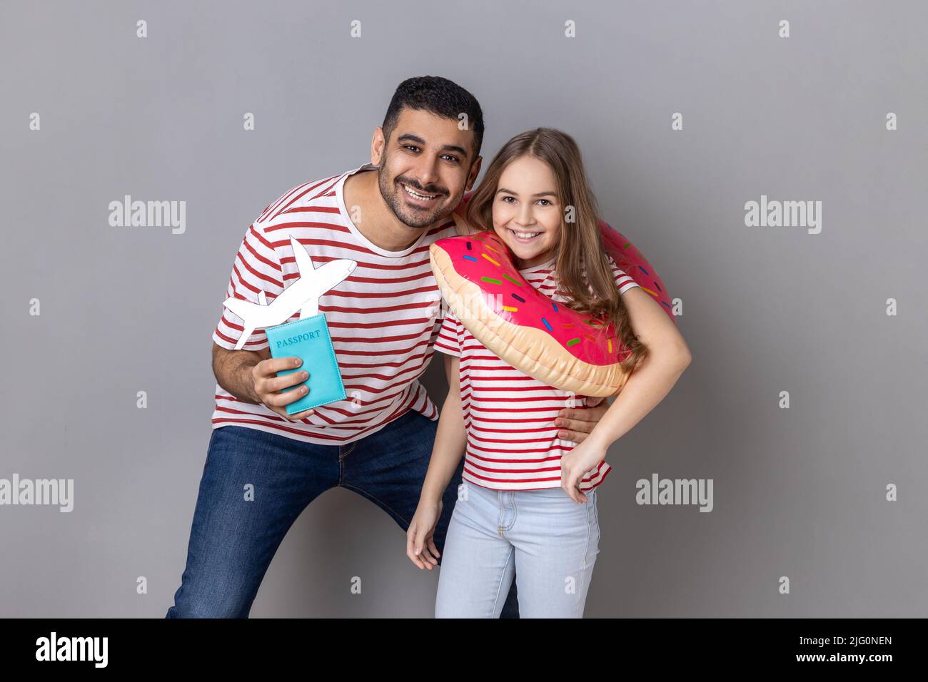 Portrait of happy dad and his daughter holding rubber ring and passport with paper plane, rejoicing their vacation abroad to the sea. Indoor studio shot isolated on gray background. Stock Photo