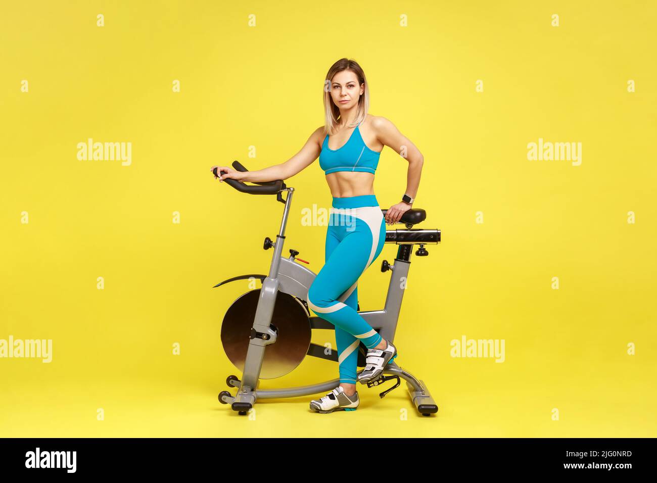 Full length portrait of attractive woman near cycle simulator and looking at camera, doing cardio workout, wearing blue sportswear. Indoor studio shot isolated on yellow background. Stock Photo