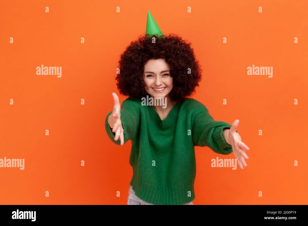 Happy satisfied woman with Afro hairstyle wearing green casual style sweater and cone, celebrating birthday party, inviting guests. Indoor studio shot isolated on orange background. Stock Photo