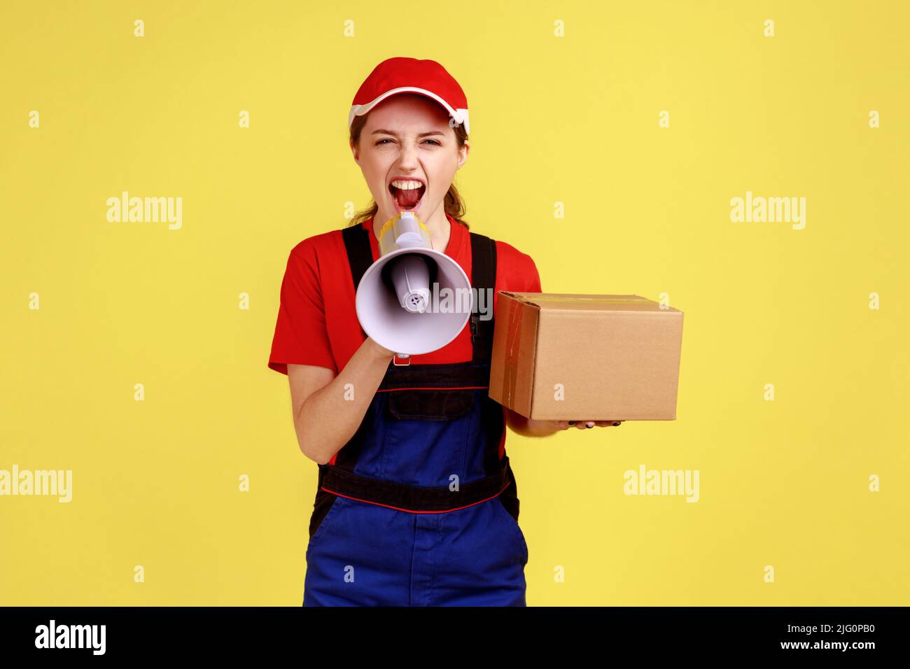 Portrait of angry courier woman holding cardboard box and screaming in megaphone, looking at camera with aggressive expression, wearing overalls. Indoor studio shot isolated on yellow background. Stock Photo