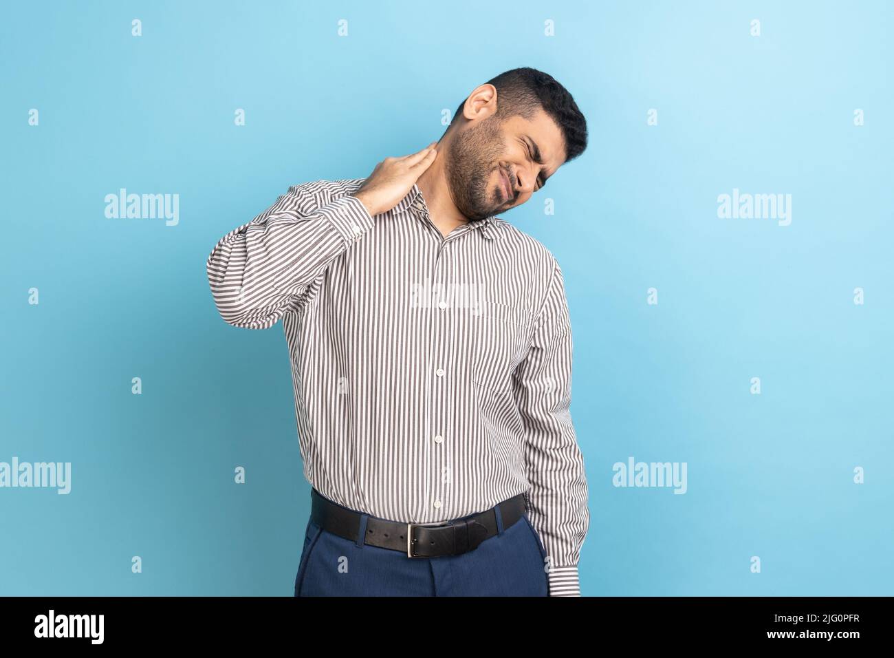 Anxious upset businessman massaging numb neck feeling pain, muscles tension, uncomfortable sleeping conditions, wearing striped shirt. Indoor studio shot isolated on blue background. Stock Photo