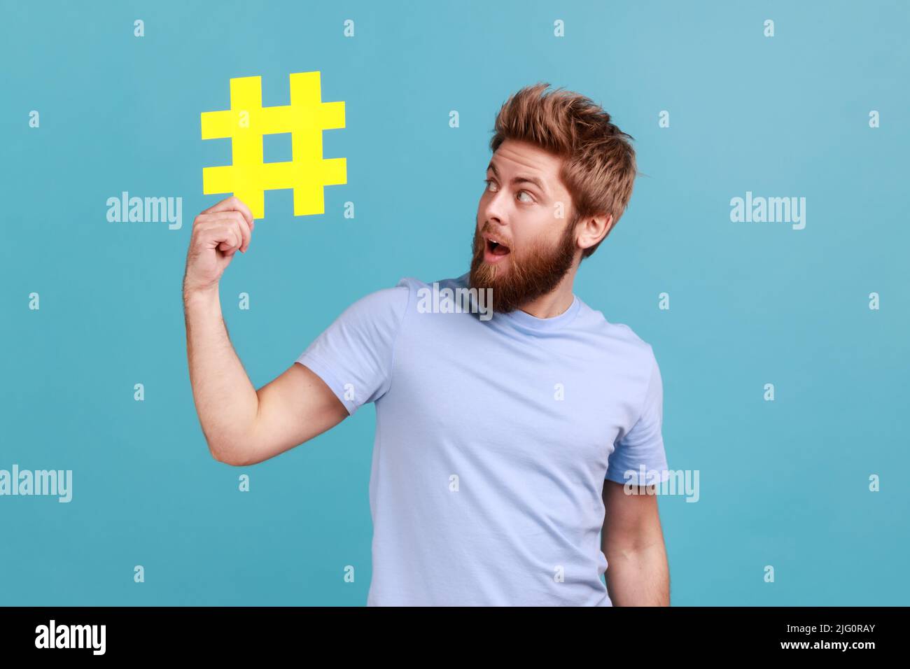 Portrait of bearded man holding yellow hashtag symbol, making important topic popular, setting trends on Internet, looking at sign with amazement. Indoor studio shot isolated on blue background. Stock Photo