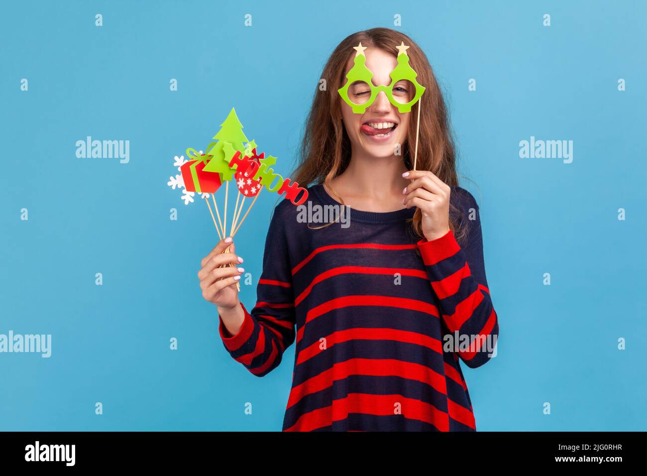 Funny positive woman wearing striped casual style sweater, holds Christmas party props, covers eyes with paper glasses, showing tongue out, festive mood. Indoor studio shot isolated on blue background Stock Photo