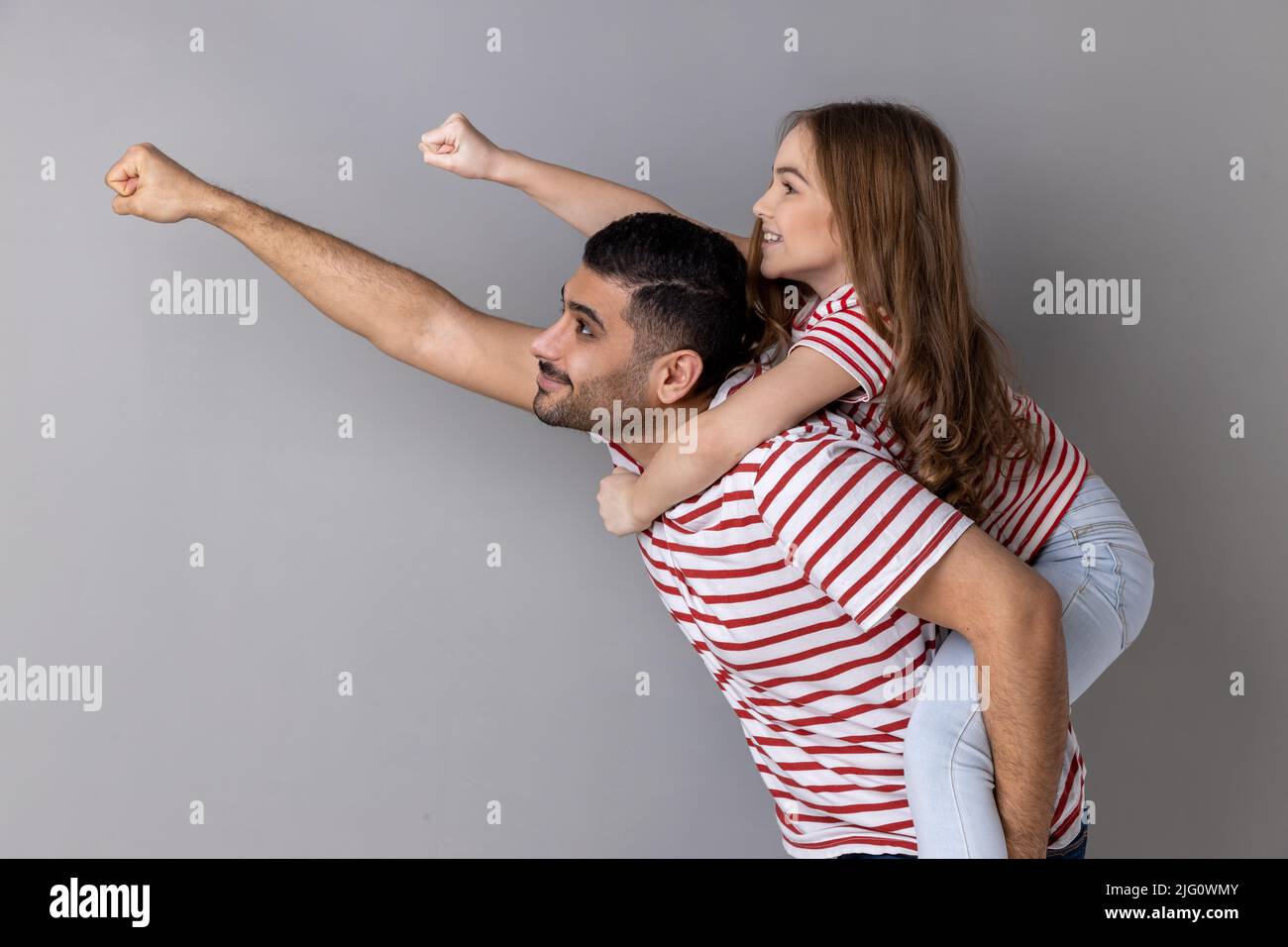 Cheerful daddy piggybacking daughter, making flying superhero hand, fist forward gesture, little girl riding fathers back, playing active game together. Indoor studio shot isolated on gray background. Stock Photo