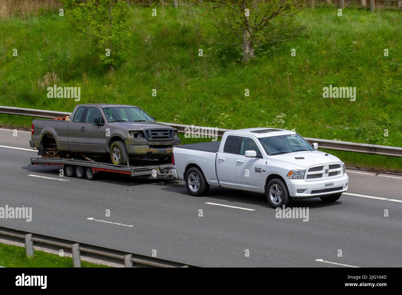 2012 white American Dodge 5654cc petrol SUV pick-up towing 3 axle trailer carrying Ford F-150 Truck wreck; driving on the M6 Motorway, Manchester, UK Stock Photo