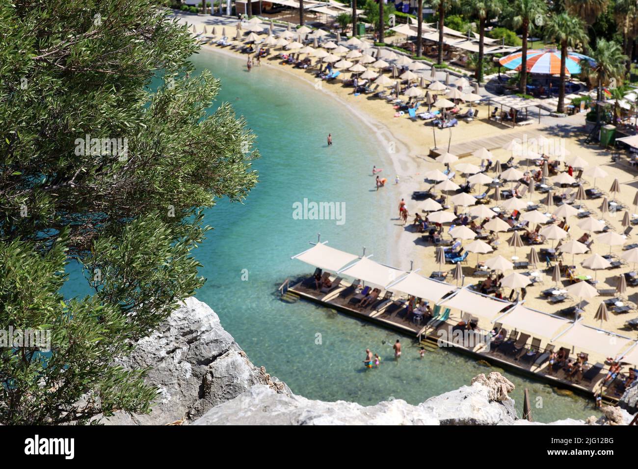 Defocused view from top of a rock to azure bay and beach with tanning and swimming people. Summer resort with parasols and lounge chairs Stock Photo