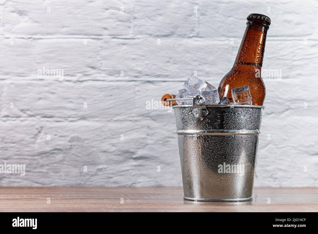 https://c8.alamy.com/comp/2JG14CF/ice-filled-metal-bucket-with-a-fresh-bottle-of-beer-capped-with-condensation-drops-on-a-wooden-table-in-front-of-a-white-wall-2JG14CF.jpg