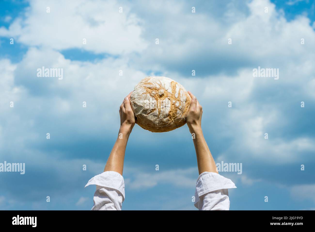 Female hands holding home baked bread loaf above her head over a blue summer sky. World food security concept. Stock Photo