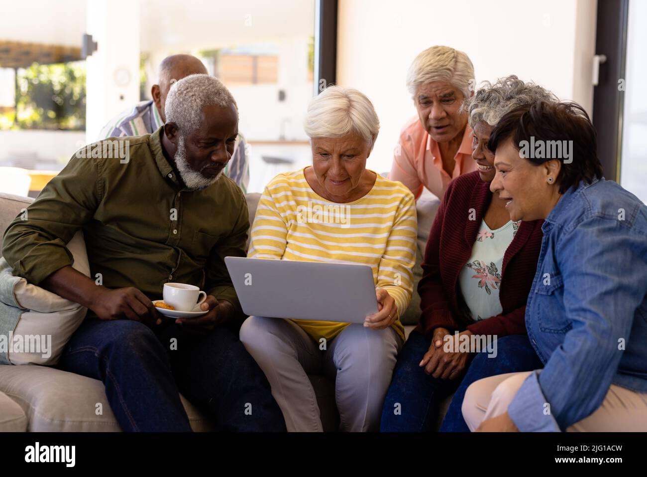 Multiracial seniors looking at female friend using laptop while sitting on sofa in nursing home Stock Photo