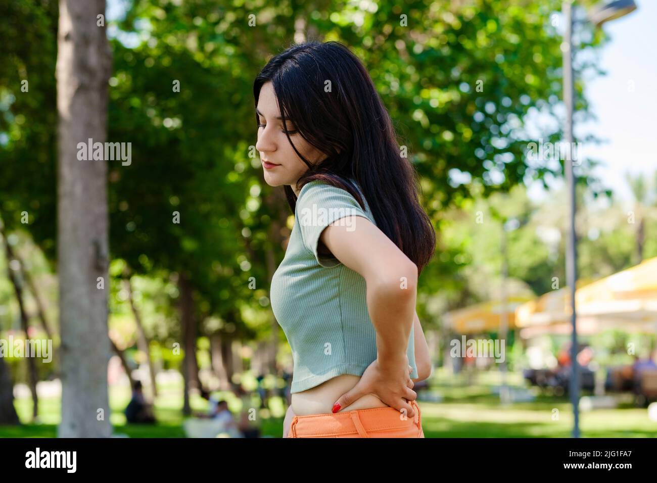 Young beautiful woman wearing turquoise tee on city park, outdoors back pain suffer from lower lumbar discomfort muscle pain. Massaging tense muscles. Stock Photo