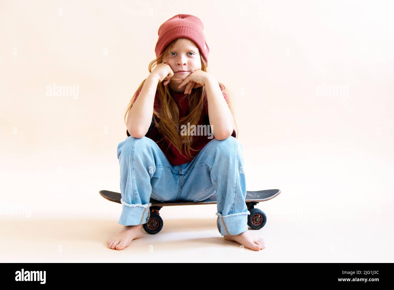 Boy with long red hair sitting on a skateboard on a light background.Front view .Hipster character. Outdoor summer fun. School background.  Stock Photo