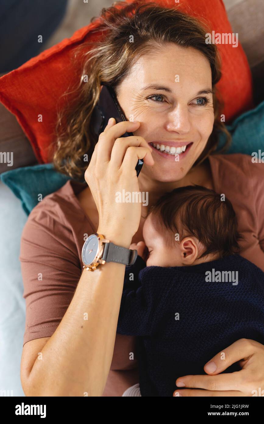 Smiling mid adult caucasian mother answering smart phone with sleeping newborn baby on couch Stock Photo