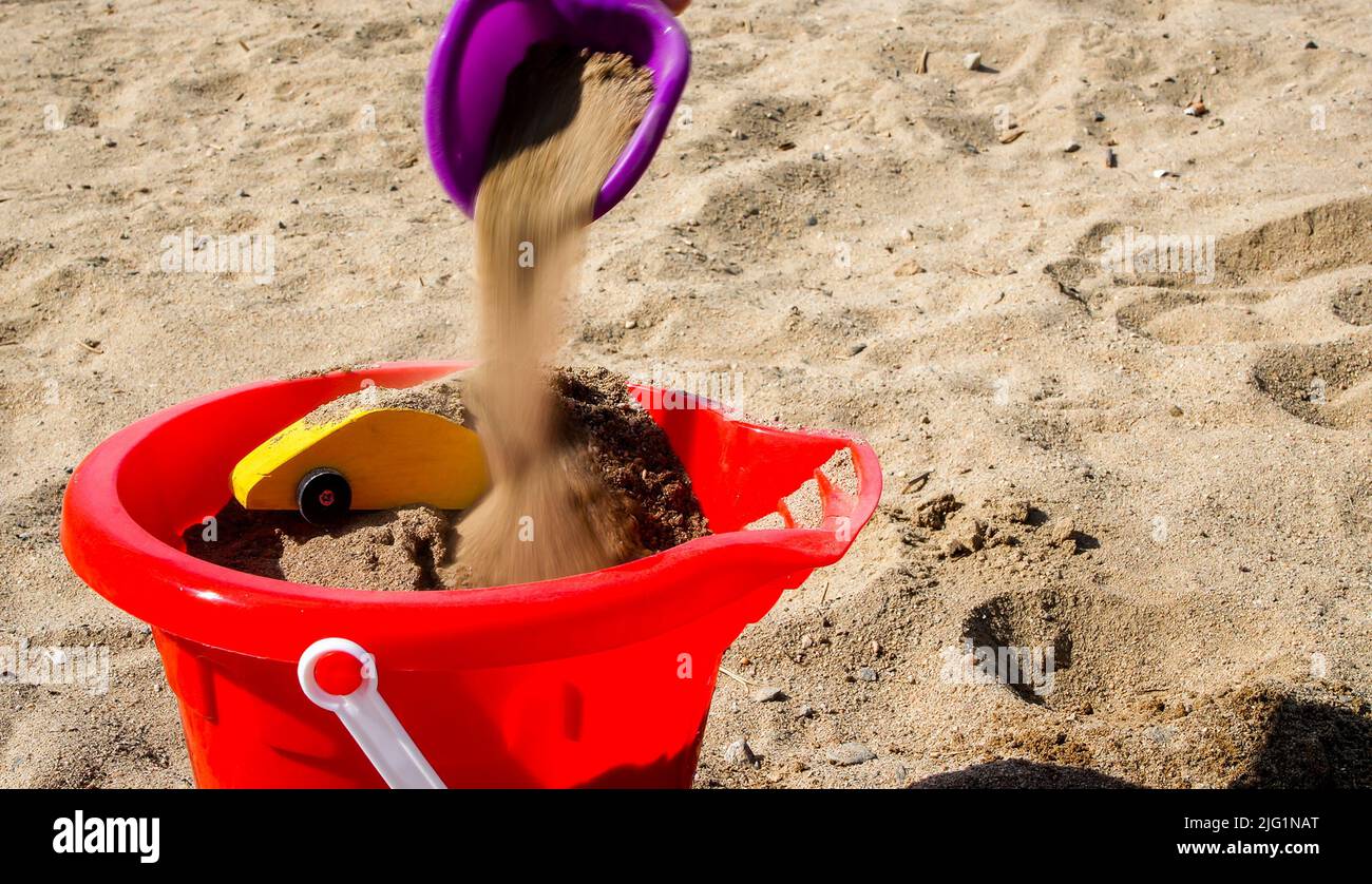 Toy shovel adding sand to yellow toy car inside red bucket on beach