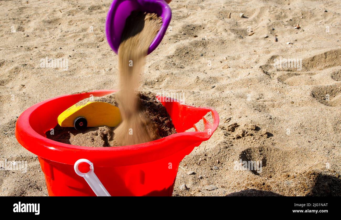 Toy shovel adding sand to yellow toy car inside red bucket on  beach Stock Photo