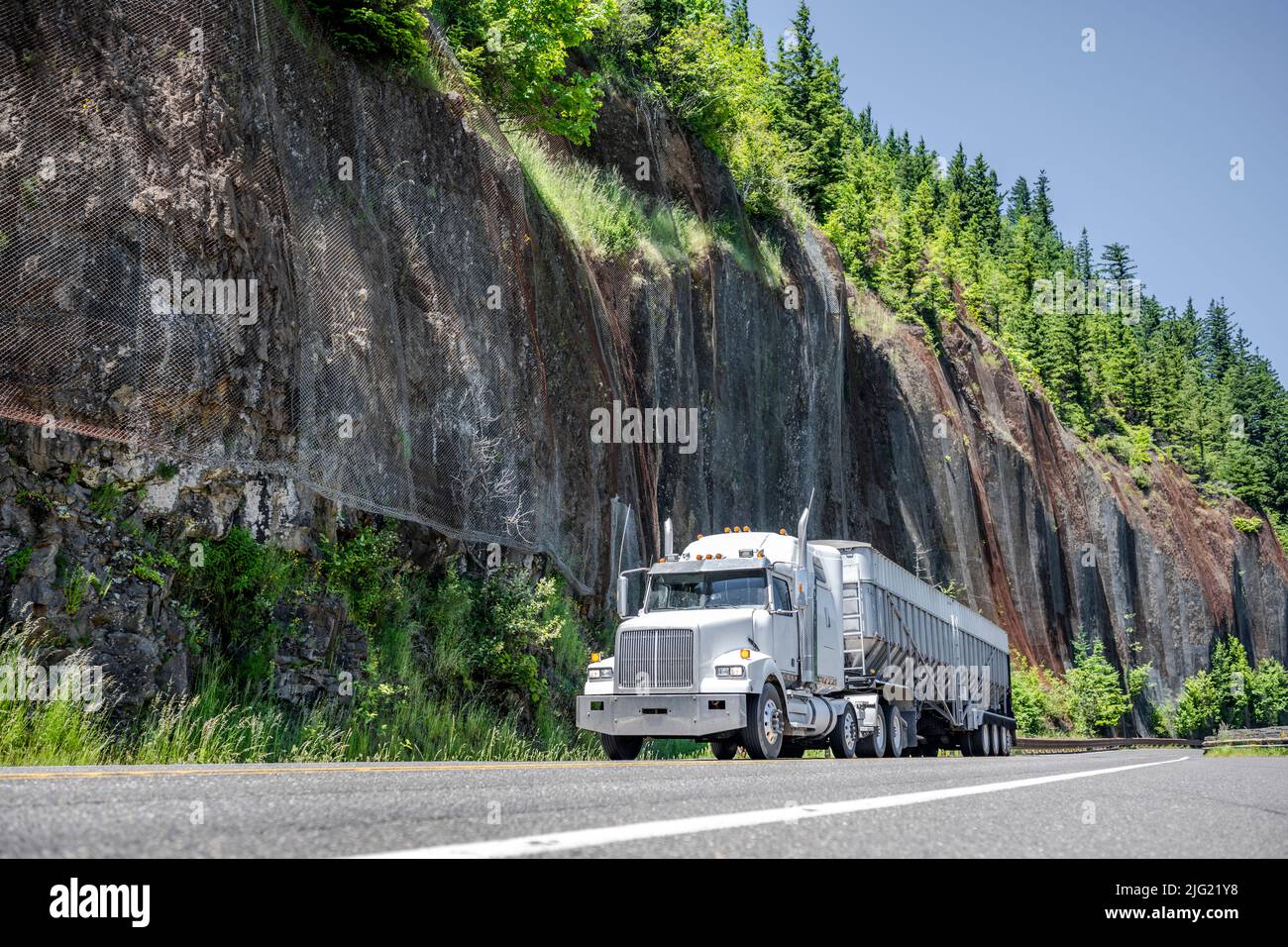 Powerful white big rig semi truck tractor transporting cargo in bulk semi trailer driving on the winding mountain narrow highway road with rock wall a Stock Photo