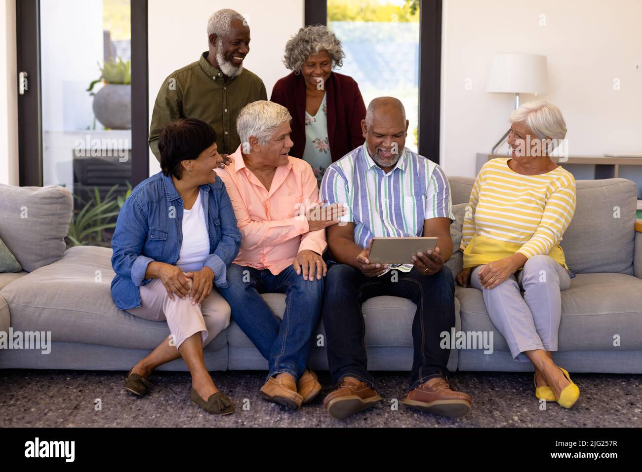 Cheerful multiracial senior friends looking at senior man using digital tablet while sitting on sofa Stock Photo