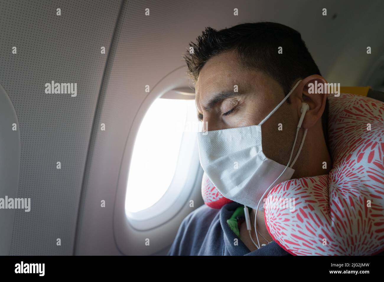 Man with face mask, earphones and neck pillow sleeping on plane window
