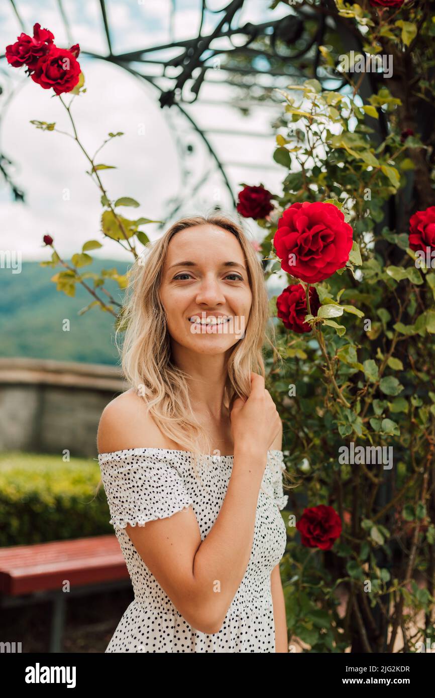 A beautiful woman stands near a bush of red roses and smiles Stock ...