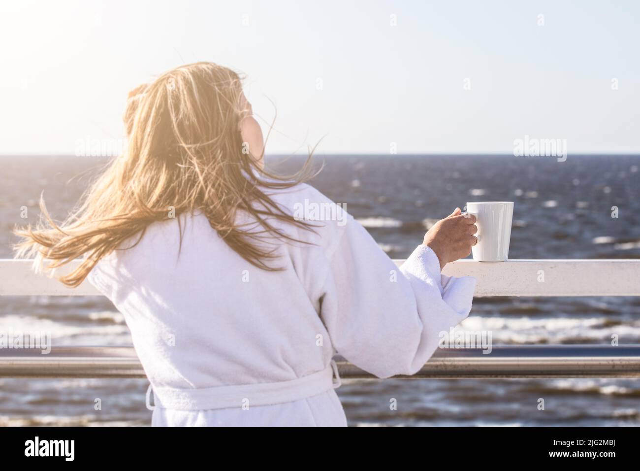 A woman in a white robe with a white mug in her hands on the terrace looks at the sea, a romantic mood.. Stock Photo