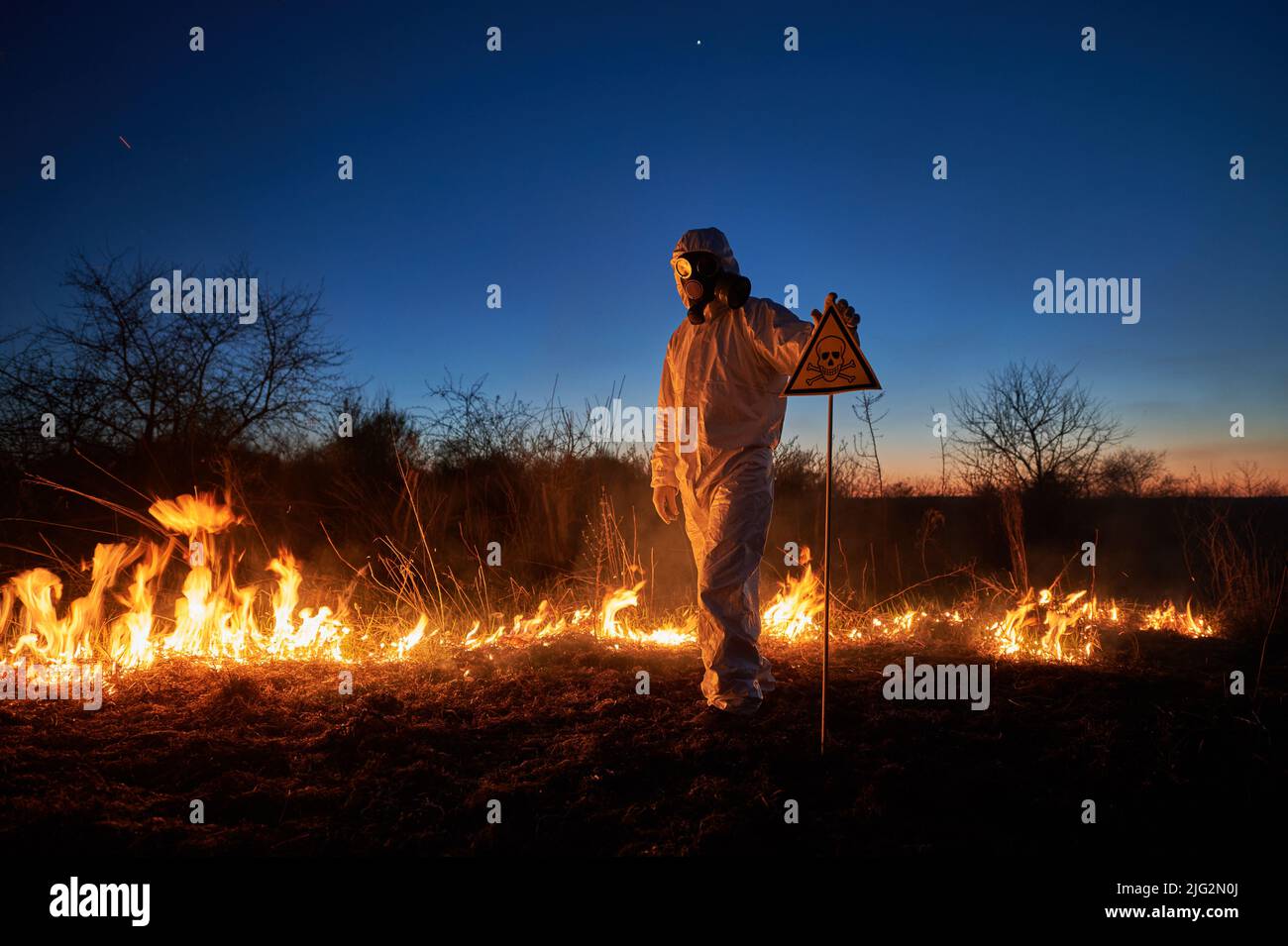 Firefighter ecologist extinguishing fire in field at night. Man in protective suit and gas mask near burning grass with smoke, holding warning sign with skull and crossbones. Natural disaster concept. Stock Photo
