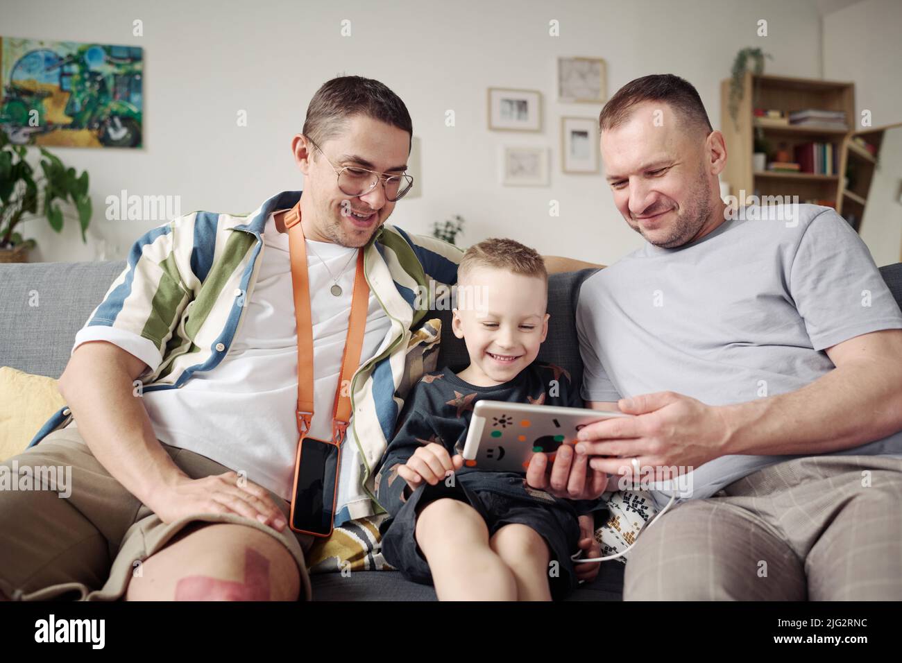 Gay couple sitting on sofa with their adopted son and watching funny video on digital tablet Stock Photo