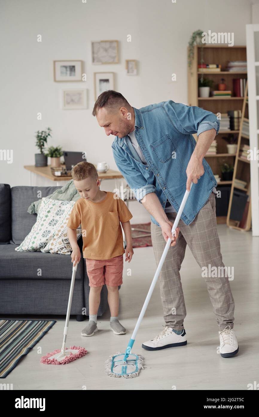 Gay father teaching his adopted child to wash floor with mop during housework in the room Stock Photo