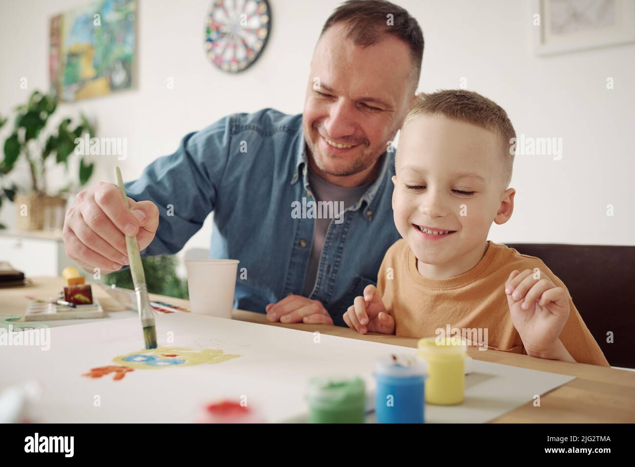 Happy father painting a picture with paintbrush together with his little son at table Stock Photo