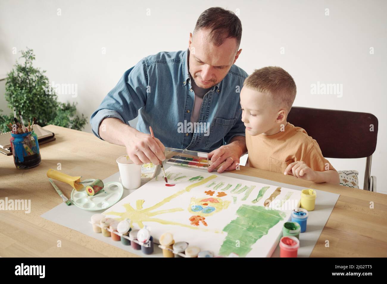Young father using paintbrush and paints to help his little son to paint a picture on paper at table Stock Photo