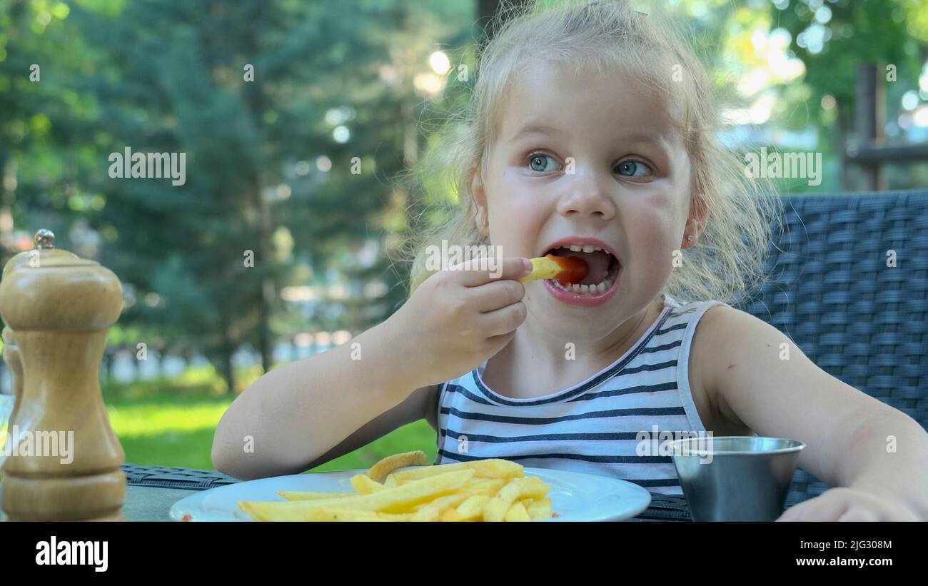 Little girl eat french fries. Close-up of blonde girl takes potato chips with her hands and tries them sitting in street cafe on the park. Stock Photo