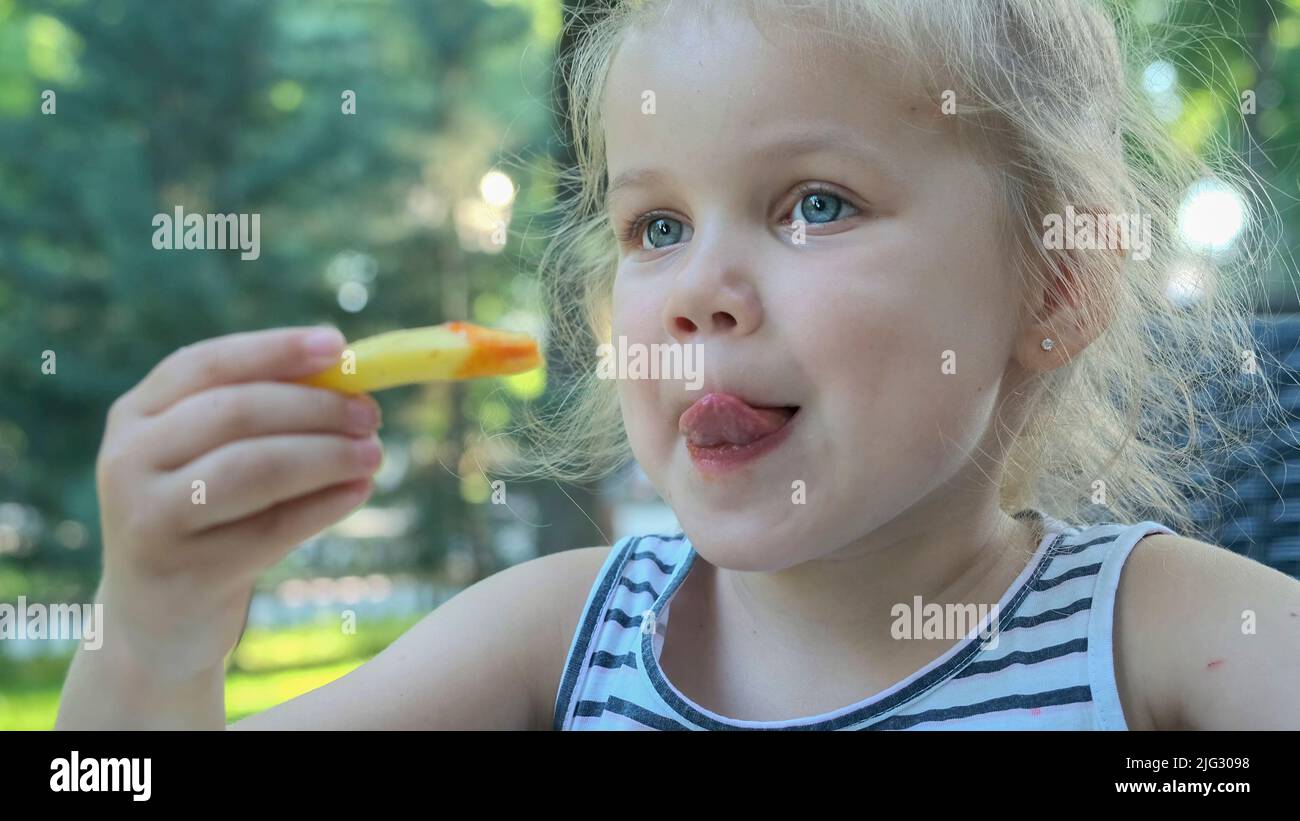 Little girl eat french fries. Close-up of blonde girl takes potato chips with her hands and tries them sitting in street cafe on the park. Stock Photo