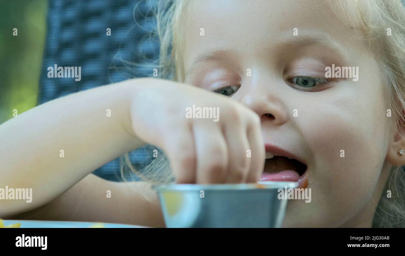 Little girl eat french fries. Close-up of blonde girl takes potato chips with her hands and tries them sitting in street cafe on the park. Stock Photo