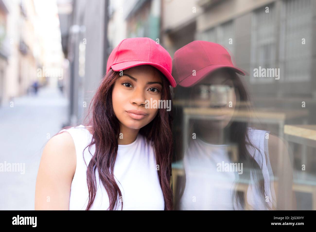 Through the glass view of stylish dressed young woman looking at camera while standing on the street in the city Stock Photo