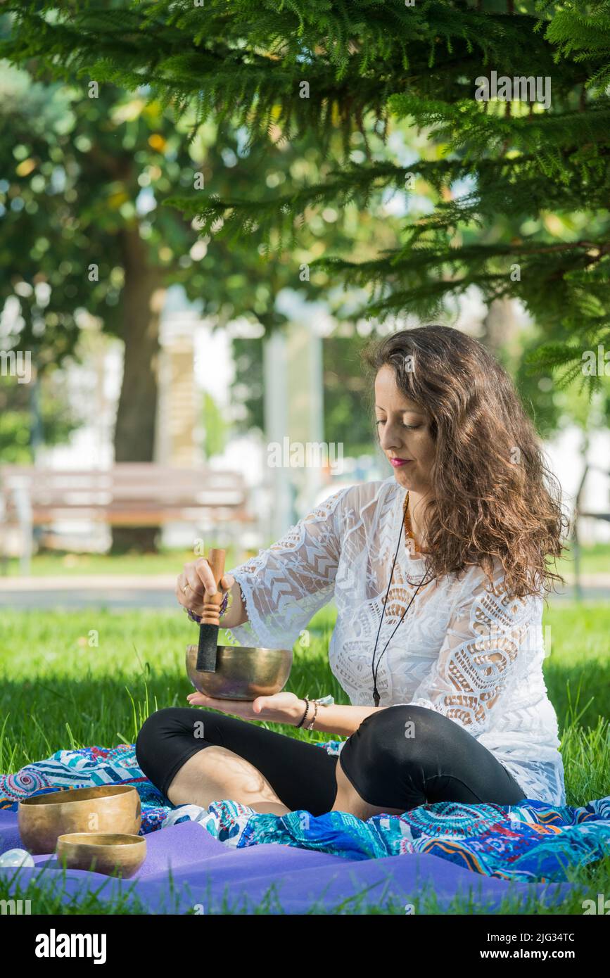 A woman, seated on yoga mats, plays a Tibetan singing bowl during a meditation and music therapy session in a park area. Stock Photo