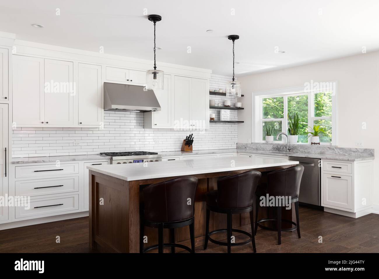 A beautiful modern farmhouse kitchen with white cabinets, a subway tile backsplash, chairs sitting at a large wood island, and marble countertops. Stock Photo