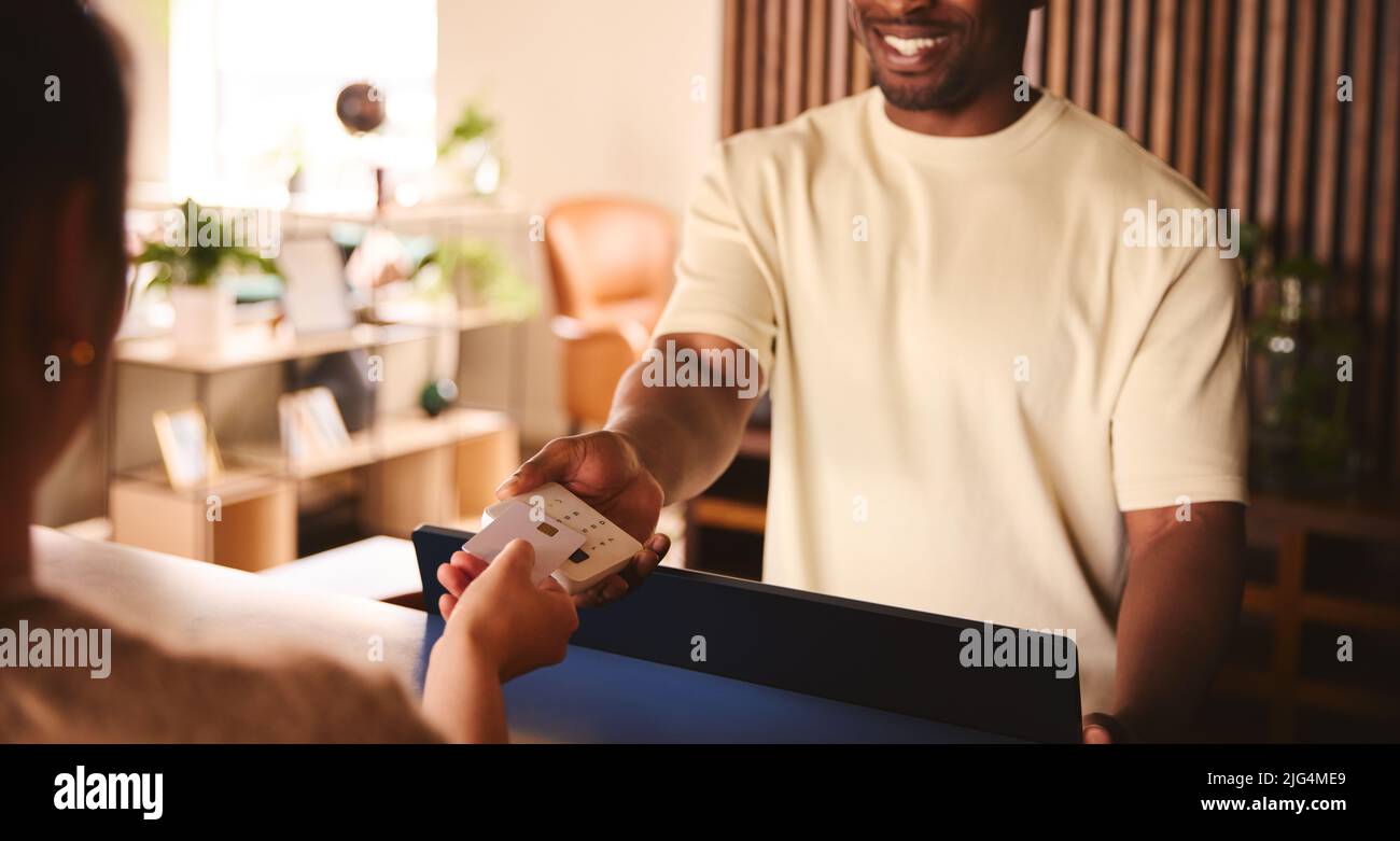 Happy Sales Assistant In Retail Shop With Customer Paying Using Contactless Payment Credit Card NFC Stock Photo