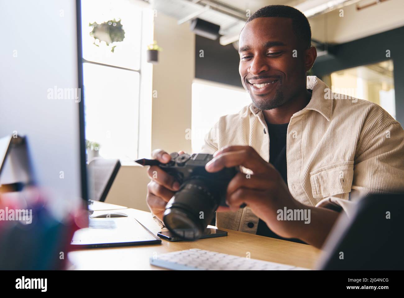 Young Black Professional Photographer Sitting At Desk Working On Computer Holding Camera Editing Pictures Stock Photo