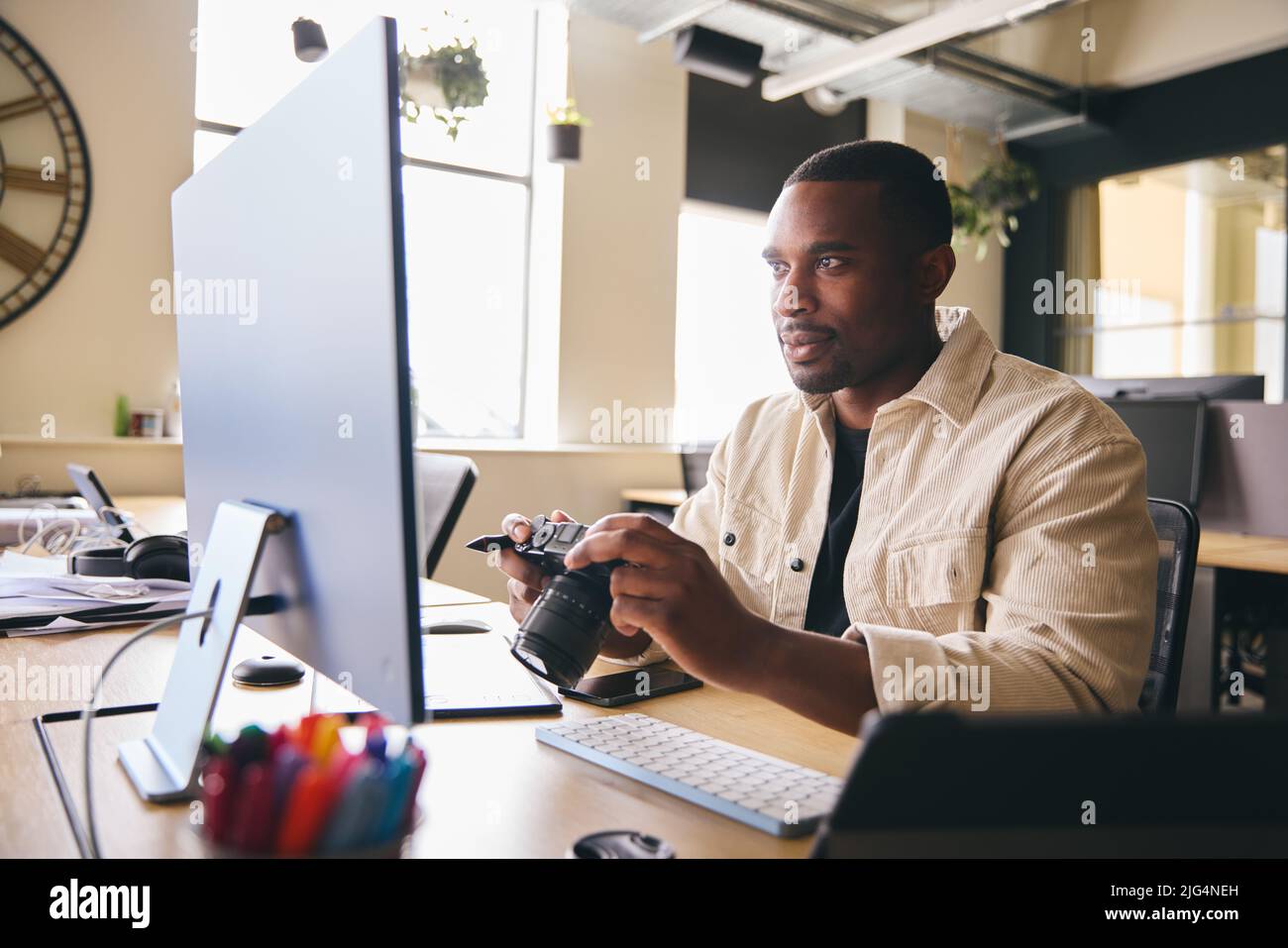 Young Black Professional Photographer Sitting At Desk Working On Computer Holding Camera Editing Pictures Stock Photo