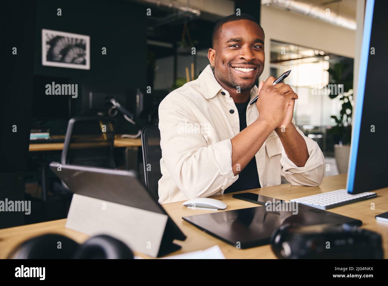 Young Black Male Advertising Marketing Or Design Creative In Modern Office Sitting At Desk Working On Computer Smiling to Camera Stock Photo