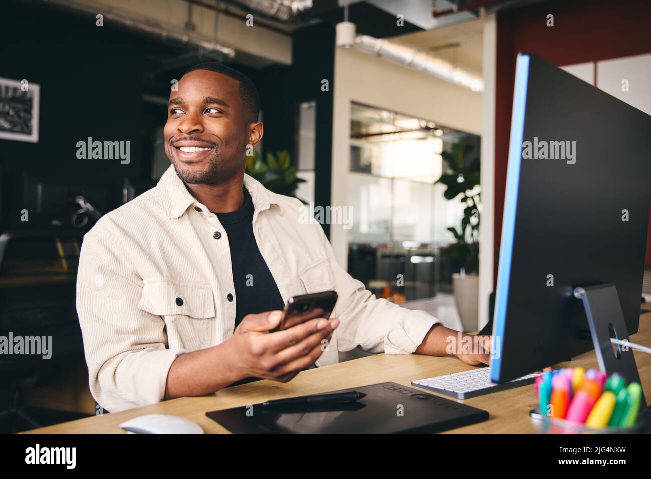 Young Black Male Smiling Advertising Marketing Or Design Creative In Modern Office Sitting At Desk Using Mobile Phone Stock Photo