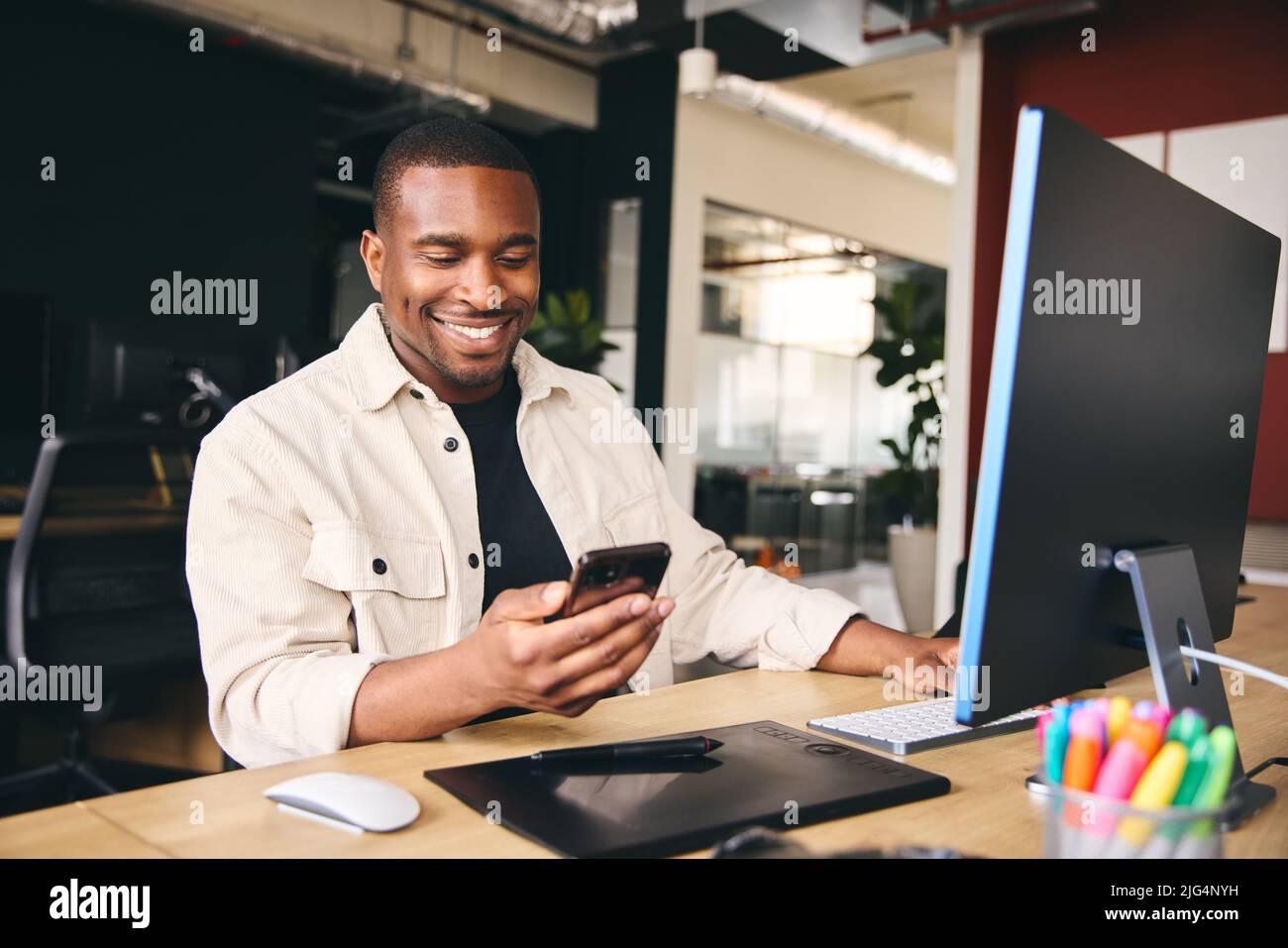 Young Black Male Advertising Marketing Or Design Creative In Modern Office Sitting At Desk Using Mobile Phone Stock Photo
