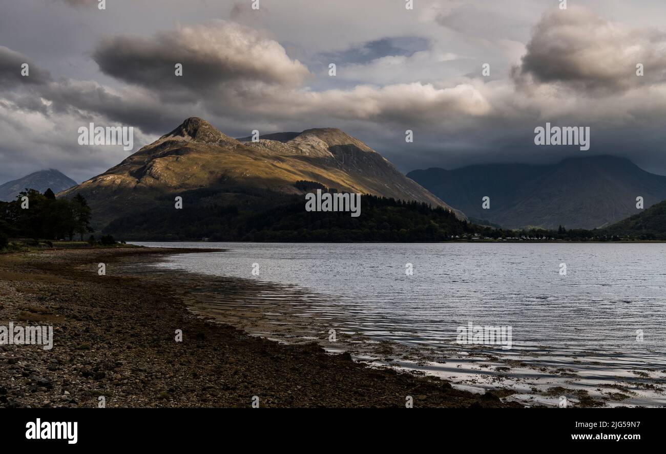 Evening view across bright water of Loch Leven to the sunlit Pap of Glencoe with the mountains at the western end of Glencoe in shadow under low cloud Stock Photo