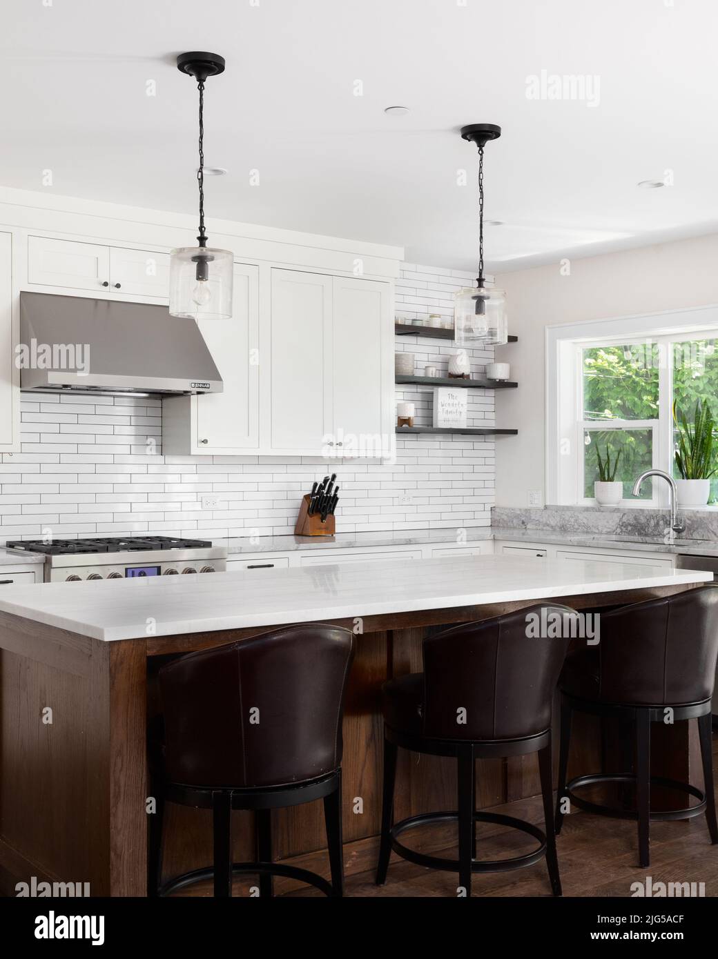 A beautiful modern farmhouse kitchen with white cabinets, a subway tile backsplash, chairs sitting at a large wood island, and marble countertops. Stock Photo