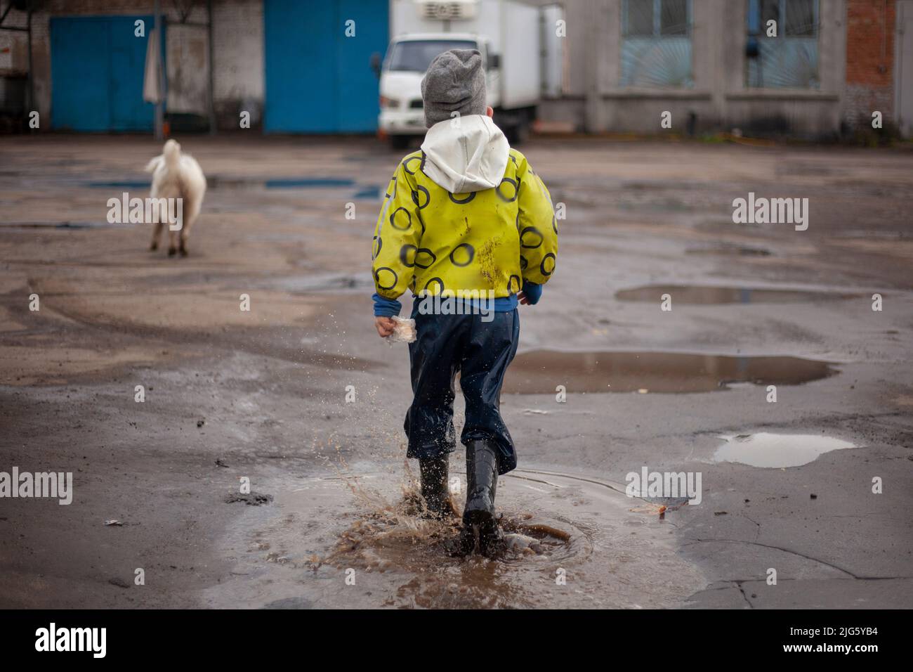 A child on a walk.  The guy in the bright jacket has fun on the street. Funny little boy. Rubber boots on the feet of the child. Stock Photo