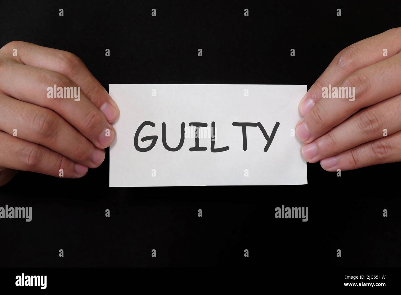 Guilty verdict concept. Criminal hands holding paper placard mug shot with written word in dark black background. Stock Photo