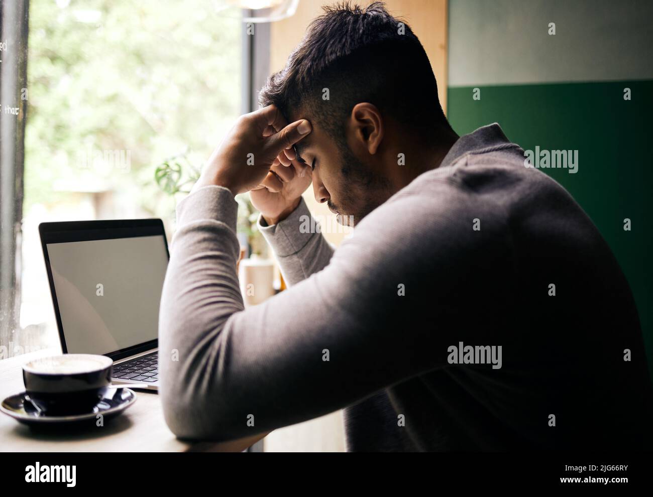 .. Stressed young mixed race man sitting at a table in a cafe drinking coffee and using a laptop alone. Unhappy hispanic businessman sitting in a Stock Photo