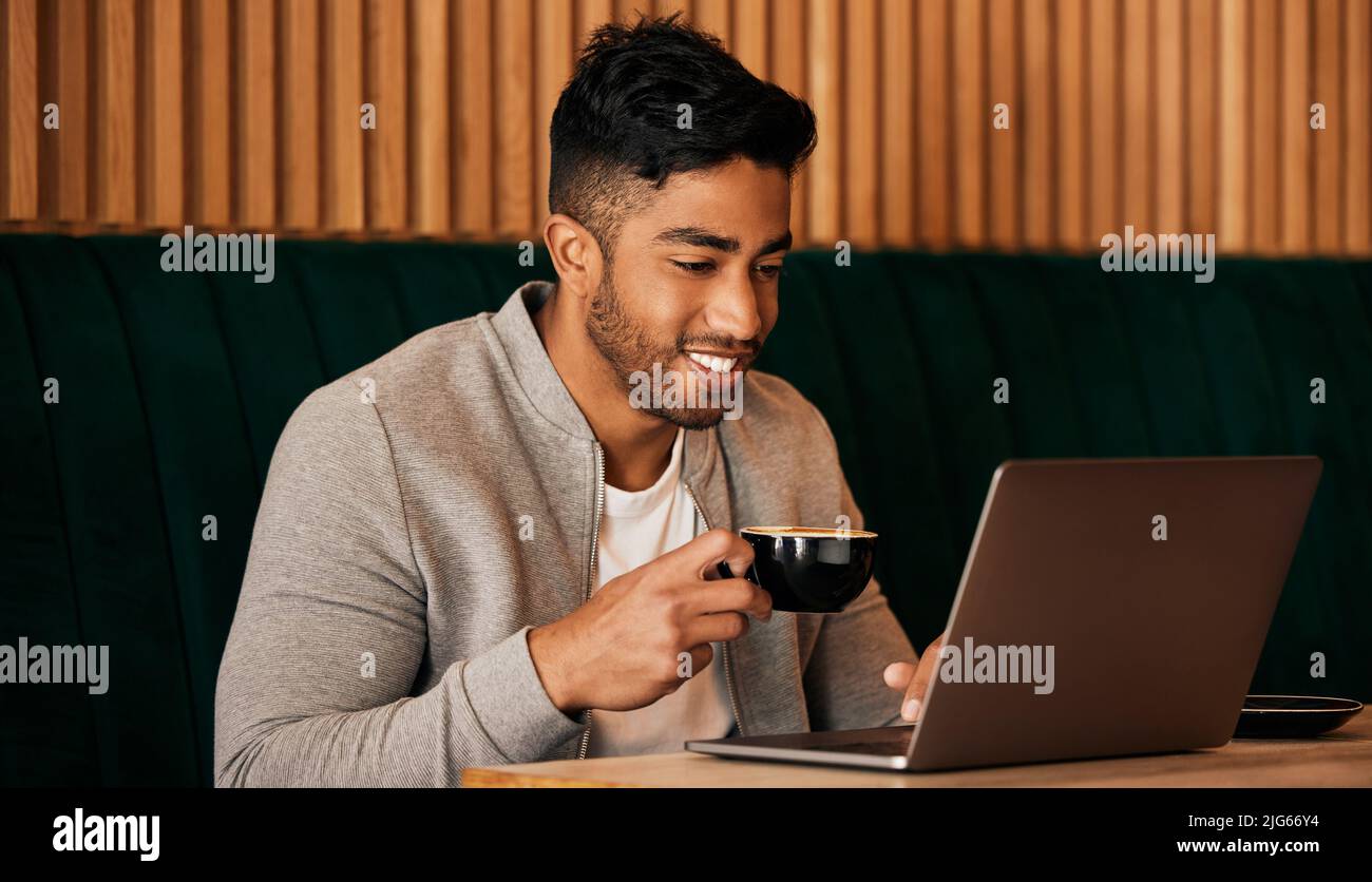 .. Happy young mixed race man sitting at a table in a cafe drinking coffee and using a laptop alone. One hispanic businessman sitting in a restaurant Stock Photo