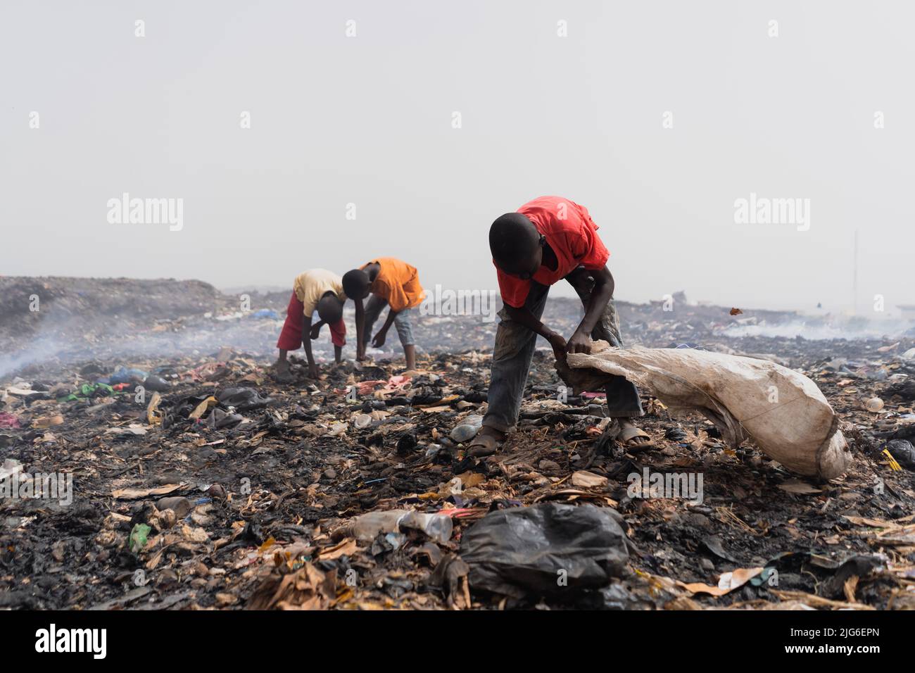 Three small African children searching for recyclable valuables at the garbage dump amidst piles of waste, dirt and smoldering smoke Stock Photo