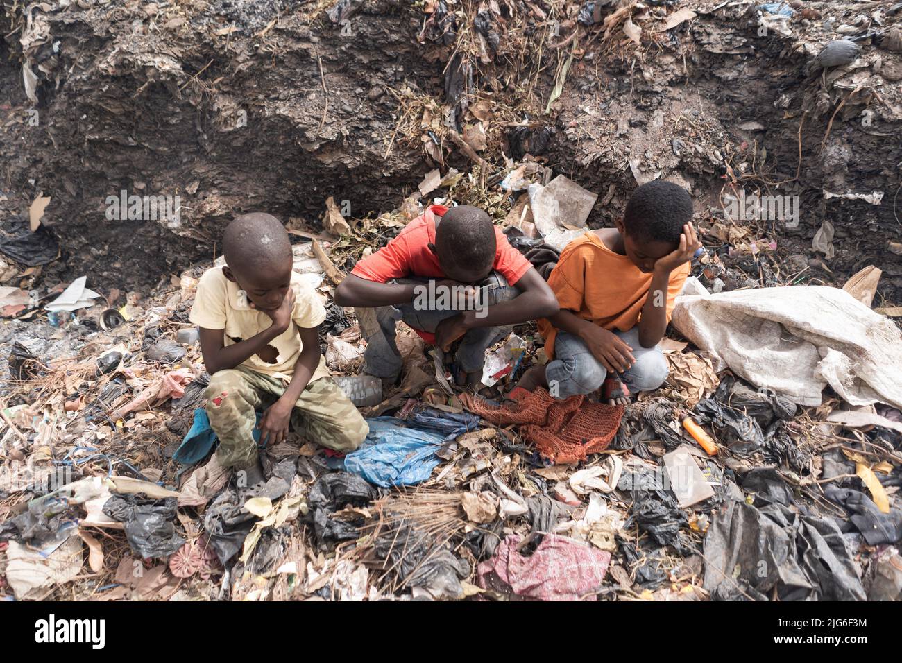 Three little African boys sitting on a landfill suffering from breathing problems, burning eyes, headaches, sore throats and the unbearable smell; Con Stock Photo